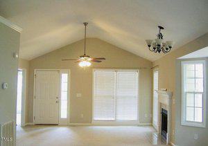 A bright, empty living room with high ceilings, neutral walls, and carpeted floors. Large windows with blinds let in natural light. There is a ceiling fan, a chandelier, and a fireplace framed by windows on the right.