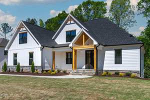 A modern house with white siding and a black roof features a welcoming front porch with wooden beams. The house is surrounded by a neatly landscaped lawn and small shrubs, set against a backdrop of green trees and a partly cloudy sky.