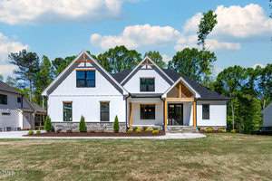 A modern farmhouse-style home with white siding, large windows, and a black roof. It features a front porch with wooden accents and stone details. The lawn is neatly manicured, with trees in the background under a blue sky with scattered clouds.