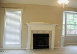 A beige living room with a white fireplace centered on the wall. To the left, a tall window with blinds, and to the right, a corner with two large windows providing natural light. The room has a ceiling fan.