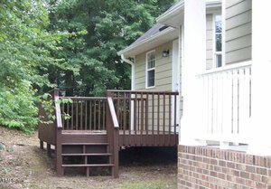 A small wooden deck with stairs, attached to a light-colored house, surrounded by greenery. The deck has a simple railing, and the house features a brick foundation and vertical siding.