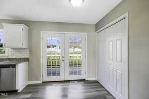 A bright room with glass French doors leading to a wooden deck outside. The room has light gray walls, white ceiling, wood-patterned floor, and a white closet on the right. There is a partial view of a kitchen with a tiled backsplash on the left.