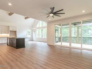 A spacious open-concept living area with wooden floors, large windows, and sliding glass doors leading to a screened porch. The room features a modern ceiling fan, a white kitchen with an island, and a chandelier near the dining space.