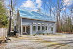 The blue-grey Bullock house at 9748 Poverty Point boasts a metal roof, numerous rectangular windows, and a gravel driveway. Surrounded by trees under a partly cloudy sky, it features a bench and planters near the entrance.