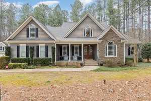 A charming two-story house on 1199 Old Still Way, with gray siding and stone accents, surrounded by tall pine trees in Wake Forest. The front porch features white columns and black wicker chairs. The well-maintained lawn and shrubs add a welcoming touch.