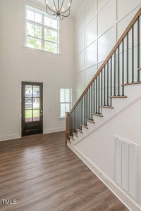 A spacious, modern entryway featuring a high ceiling and large window. The space includes a dark wooden front door and a staircase with wooden steps and black railings. The floor has a light wood finish, and the walls have a paneled design.