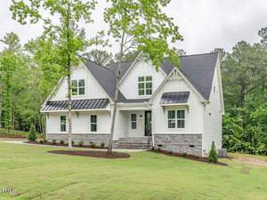A modern white house with a dark roof features a manicured lawn and young trees in the front yard. It has large windows and a stone foundation, set against a backdrop of tall trees under a cloudy sky.
