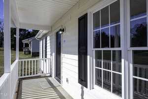 A close-up view of a house exterior showcasing a white porch with railings and a black front door. The house features large windows reflecting a clear blue sky. In the background, another house and green lawn are visible.