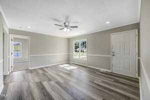 A bright, empty room with light gray walls and wood flooring. Features include a ceiling fan, a large window letting in natural light, and a white door. The room leads to another space beyond an open doorway.