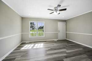 A bright, empty room with light wood flooring, neutral-colored walls, and a ceiling fan. Natural light streams through a large window and a door with a small window. The view outside shows greenery and a clear sky.