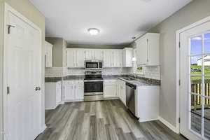 A bright kitchen with white cabinets, stainless steel appliances, and light wood flooring. The walls are light gray, and there is a door leading to a wooden deck. A window above the sink lets in natural light.