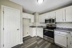 Modern kitchen with white cabinets, stainless steel appliances, and gray countertops. A white subway tile backsplash lines the walls, and light wood flooring covers the floor. A closed door is on the left.