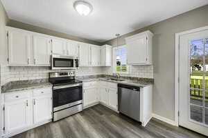 A modern kitchen with white cabinets, stainless steel appliances, and gray countertops. It features a stove, microwave, and dishwasher, with a tile backsplash. A window and sliding glass door provide natural light, overlooking a green backyard.