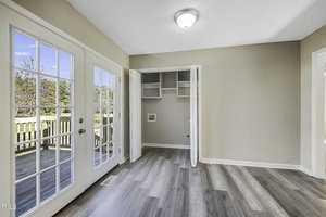 A bright room with light gray wood flooring, beige walls, and a double glass door leading to a wooden deck. An open closet with shelves is visible. Sunlight streams through the doors, illuminating the space.