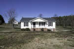 A small, white, single-story house at 110 Clubview Place, complete with a charming front porch and brick steps, sits on a grassy lot in Oxford. Sparse trees are scattered around, and the sky is clear and blue in the background.