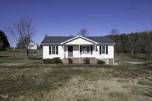 A small, white, single-story house at 110 Clubview Place, complete with a charming front porch and brick steps, sits on a grassy lot in Oxford. Sparse trees are scattered around, and the sky is clear and blue in the background.