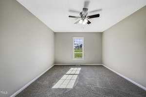 An empty room with beige walls and gray carpet, featuring a central window with a scenic view. Sunlight streams through the window, casting shadows on the floor. A ceiling fan with lights is mounted above.