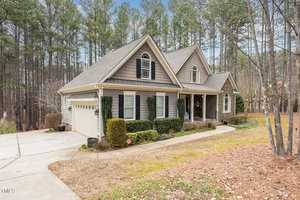 A suburban house with gray siding and a gable roof, surrounded by tall pine trees. The front has a two-car garage, white trim, dark shutters, and landscaped bushes. A concrete driveway leads up to the entrance, and the lawn shows some dry patches.