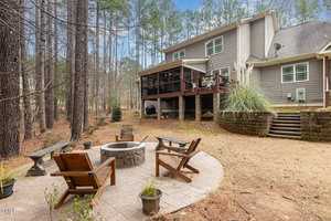 A backyard with a stone fire pit surrounded by four wooden chairs on a patio. Behind it, a multi-story house with an elevated deck and stairs. Tall trees line the background, creating a serene forest setting.