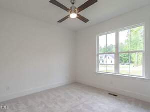A bright, empty room with white walls and a large window showing a view of a suburban neighborhood. The room features a gray carpet and a ceiling fan with lights. Natural light fills the room.