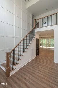 A bright entryway features a wooden staircase with black metal railings, leading to an upper level. The room is spacious with high, paneled walls and wood flooring. Large windows in the background illuminate the space with natural light.