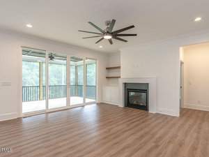 A spacious living room with wooden floors, a modern ceiling fan, and a fireplace. Glass doors open to a wooden deck with a view of greenery. Shelves are built into the wall beside the fireplace. The room is well-lit with recessed lighting.