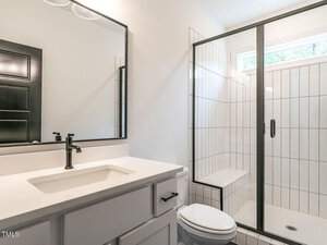 Modern bathroom with a white vanity, black faucet, and rectangular mirror. Features a white-tiled shower with a black-framed glass door and a small window above for natural light. White walls and a sleek design complete the space.