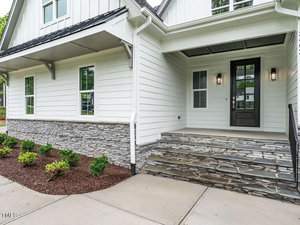A modern house entrance with stone steps and a dark wooden door. The exterior features white siding with stone accents. Small shrubs are planted alongside the walkway leading to the entrance.