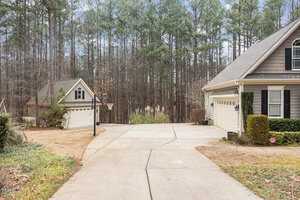 A driveway leads to a house and a detached garage, both with gray siding and white trim. A basketball hoop is positioned near the garage. In the background, a dense forest of tall pine trees is visible. The area is surrounded by manicured lawns.