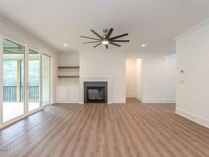 A bright, modern living room with wood flooring, a ceiling fan, and built-in shelves. The room features a fireplace with a white mantel. Large sliding glass doors lead to an outdoor deck, letting in natural light.