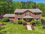 The charming two-story red brick house at 4057 Chewning Road, Oxford boasts a neatly manicured lawn and hedges. With two windows on each side of the central entrance and a chimney on the right, it sits against a backdrop of lush green trees under a clear blue sky.