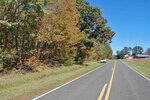 The rural stretch of 0 Goshen Road with its double yellow line extends into the distance. Trees with autumn foliage line the left side, and a white car rests on the right. A house with a red roof is visible further down, under Oxford's clear blue sky.