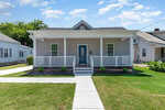 A small, single-story house at 119 Sycamore Street features a gray exterior with a charming blue front door. The Oxford-style covered porch boasts white railings, while a walkway meanders through the lush grassy lawn beneath a clear sky with a few clouds.