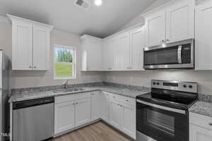 Modern kitchen with white cabinetry, granite countertops, and stainless steel appliances, including a dishwasher, microwave, and oven. A small window lets in natural light, and light wood flooring complements the space's clean, bright appearance.
