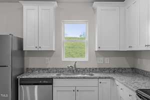 A modern kitchen with white cabinets and a speckled granite countertop. The kitchen features a stainless steel dishwasher and fridge. A small window above the sink provides a view of a grassy landscape outside.