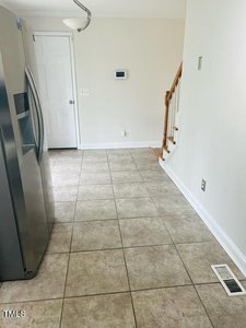 A kitchen area with tiled flooring, featuring a stainless steel refrigerator on the left. A white door, thermostat, and wooden staircase are visible in the background. The walls are painted off-white.