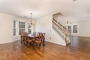 A dining area with a wooden table and chairs next to a staircase leading upstairs. The room has hardwood floors and large windows, offering natural light. A chandelier hangs above the table, and a ceiling fan is visible in the adjacent room.