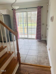 A bright kitchen area with tiled flooring, featuring a sliding glass door with pink patterned curtains leading to an outdoor space. A staircase with wooden steps is on the left, and a part of the fridge is visible to the left.