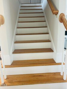 A wooden staircase with light brown steps and white risers leads upward. The stairs are bordered by white railings with wooden handrails. The walls alongside the stairs are painted white, creating a clean and minimalistic look.