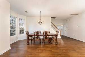 Dining area with a wooden table and six chairs. A decorative chandelier hangs above the table. The room features large windows on the left and hardwood flooring. A staircase and doorways are visible in the background. Bright, neutral walls.