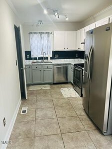 A modern kitchen with white cabinets, a marble countertop, stainless steel appliances, including a refrigerator and stove, and a double sink beneath a window with a sheer curtain. Tile flooring completes the space.