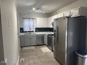 A modern kitchen with stainless steel appliances, including a refrigerator, stove, and dishwasher. White cabinets and marble countertops complement the tiled floor. A window with a patterned curtain is above the sink, allowing natural light in.