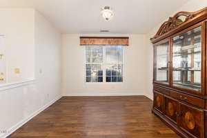 An empty room with hardwood floors and a large window. A decorative china cabinet with glass doors stands on the right. The walls are white, and a small ceiling light fixture provides illumination.