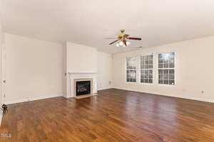Empty living room with wooden floors, white walls, and large windows. A white fireplace is centered against one wall, and a ceiling fan with lights is overhead. Natural light brightens the space.