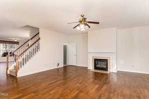 A spacious, empty living room with hardwood floors and a ceiling fan. There is a fireplace with a white mantel on the right wall. A staircase with a wooden railing leads upstairs, and an open doorway leads to another room.