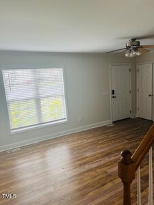 A bright, empty room with light-colored walls and laminate flooring. A large window with blinds lets in natural light. A ceiling fan is installed on the ceiling, and there is a white front door with a side closet door visible.