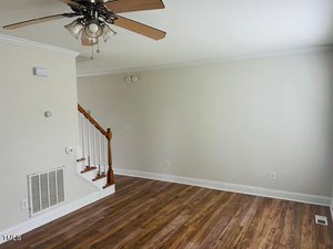 A bright living room with light-colored walls, wooden flooring, and a ceiling fan with lights. A wooden staircase with white railings is visible in the corner. There are a few electrical outlets and vents on the walls.