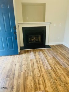 A room with light wood flooring and a white fireplace with a black interior. A blue door is partially open, revealing the wooden floor's texture. The walls are painted a light color, and the room appears empty and well-lit.