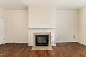 A cozy room with beige walls features a centered fireplace framed by white trim. The floor is dark wood, and there is a closed door to the left and an electrical outlet on the right wall.