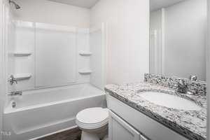 A clean, modern bathroom with white walls and fixtures. It features a bathtub with a shower, a toilet, and a vanity with a speckled granite countertop and a sink. A mirror is mounted above the vanity. The flooring is wood-patterned.